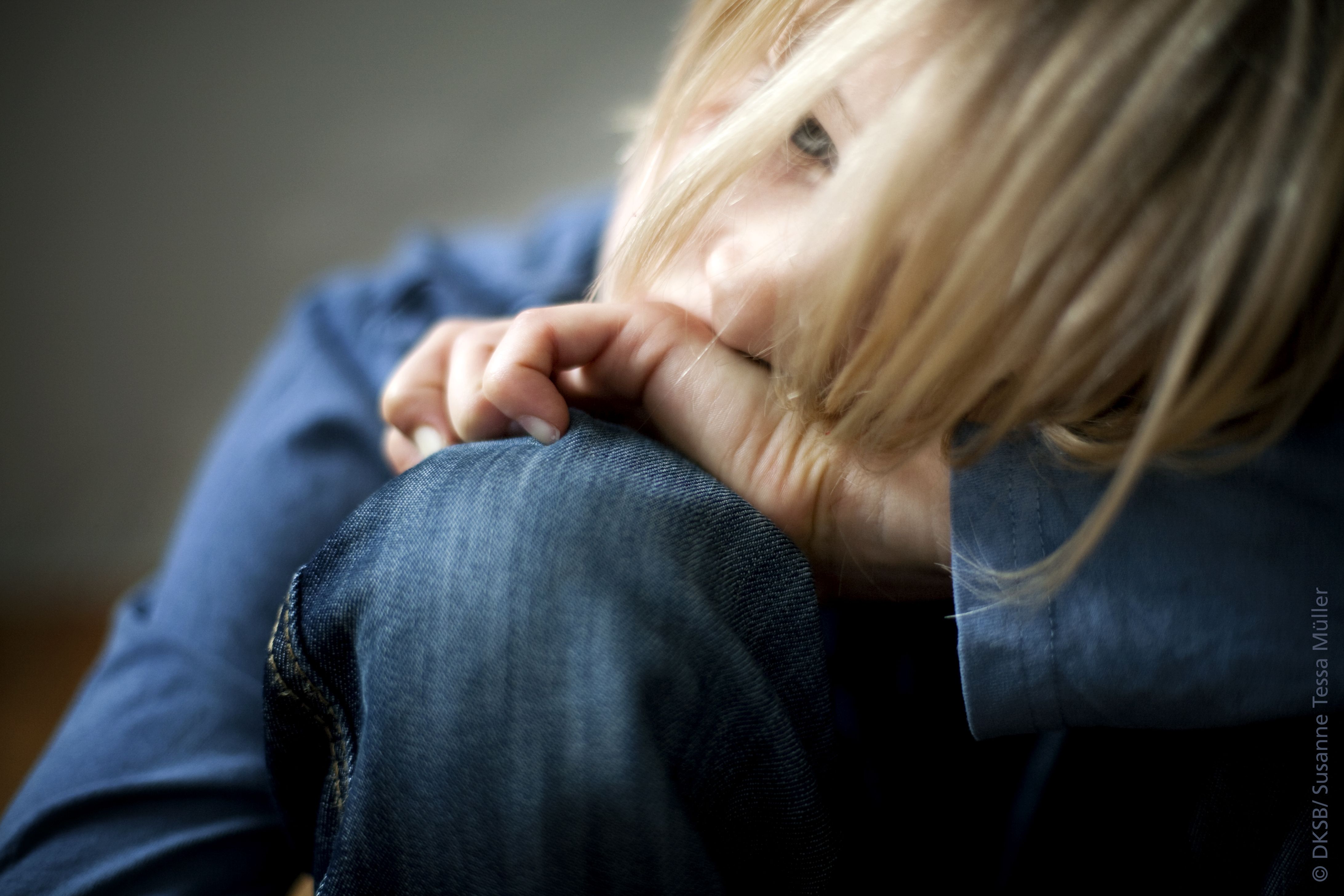 Ein nachdenkliches Kind sitzt auf dem Boden und umarmt ein Knie, während es mit langen Haaren spielt.