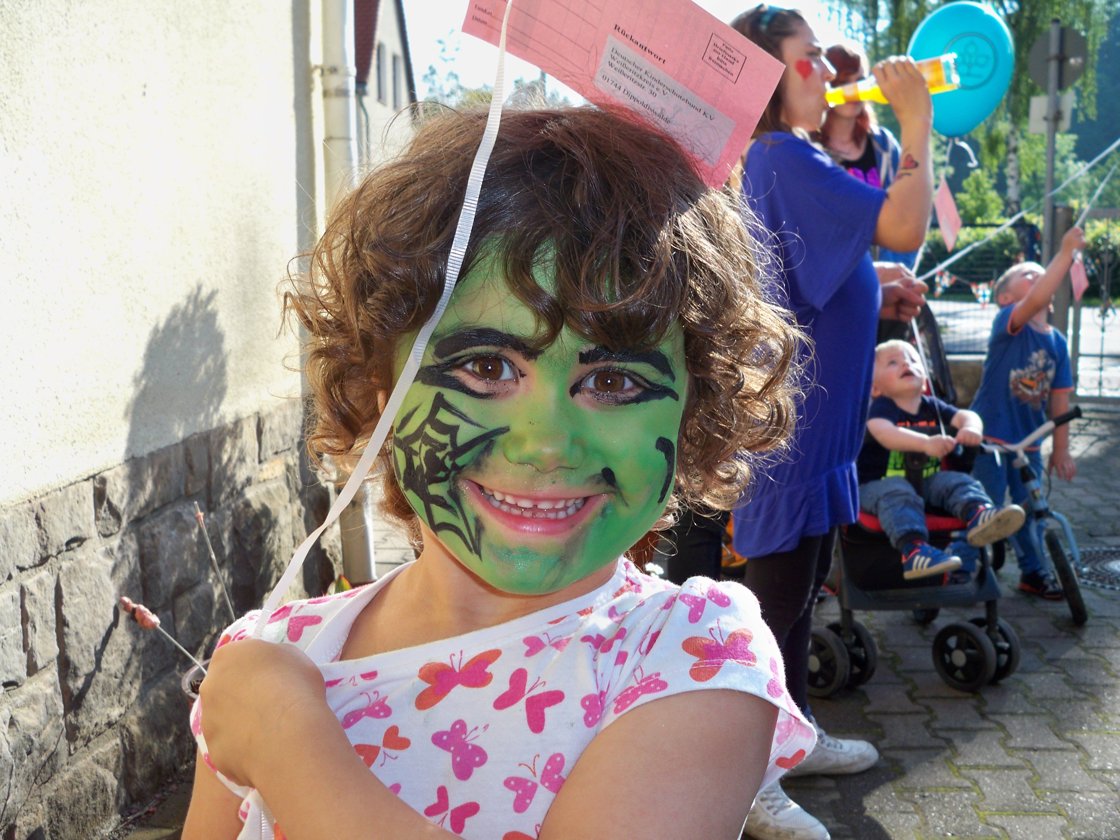 Ein lächelndes Kind mit grünem Gesicht und buntem Oberteil hält einen Luftballon und genießt eine fröhliche Feier.