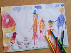Eine bunte Kinderzeichnung zeigt eine Familie vor einem Haus mit Tieren und bunten Buntstiften daneben.
