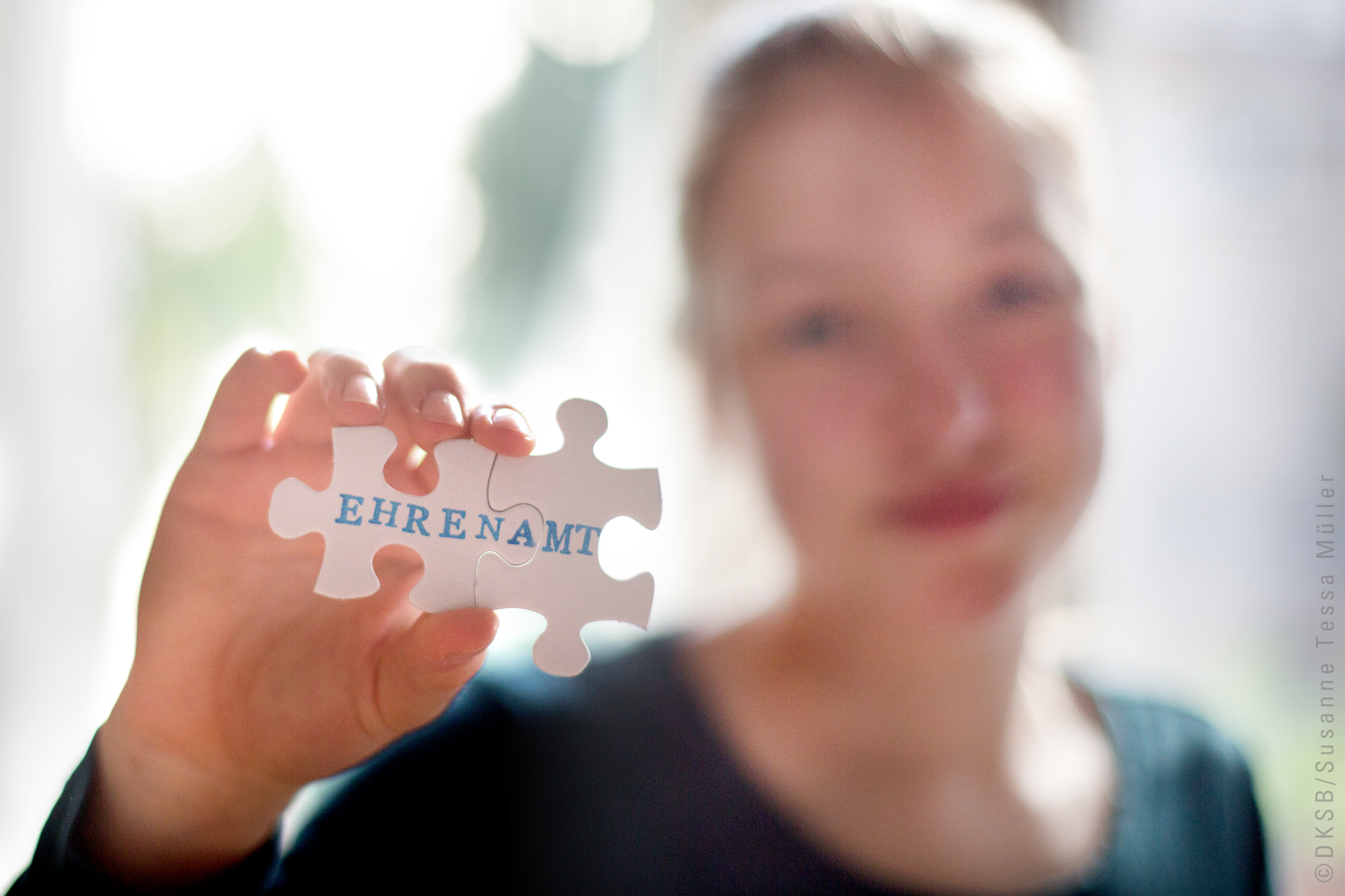 Ein Mädchen hält ein Puzzlestück mit der Aufschrift "EHRENAMT" in die Kamera und lächelt dabei freundlich.